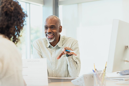 The mature adult male banker smiles and gestures while speaking to an unrecognizable female customer.